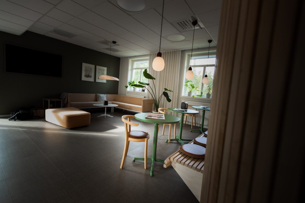 Interior image of a break room. A sofa is placed in the corner. Above the sofa hang two framed pictures. To the right in the image, three tables are lined up. Closest to the tables is a wooden bench.