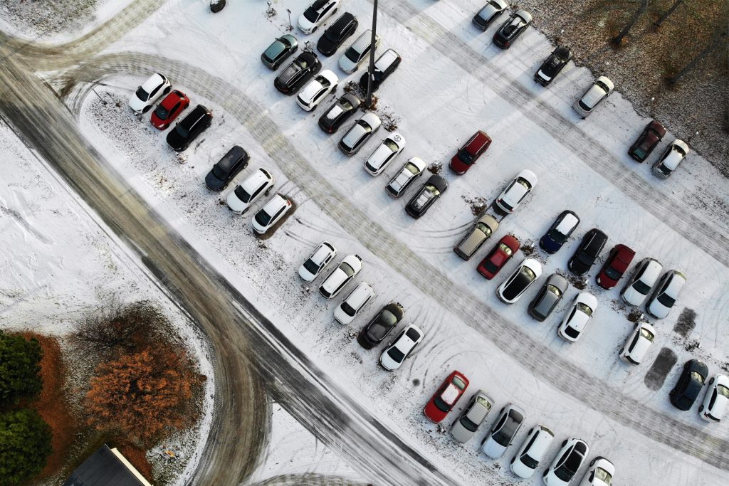 Drönarbild av en snötäckt parkeringsplats sedd ovanifrån. Flera däckspår syns i snön, vilket skapar mönster av cirklar och linjer.