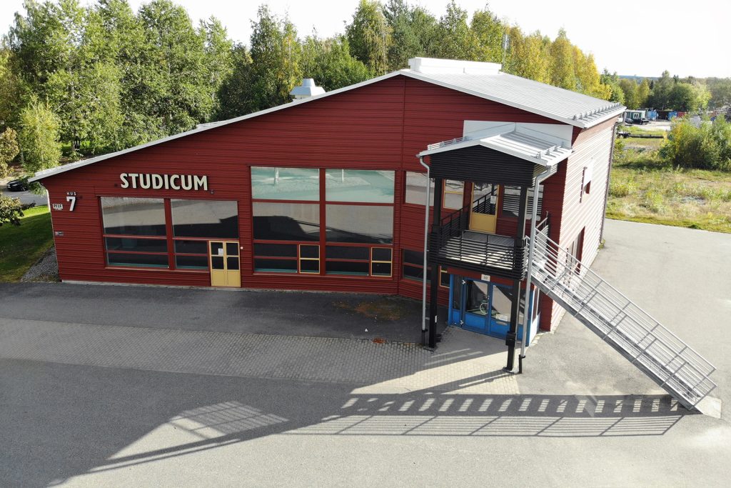 Drönarbild av ett rött trähus med lutande tak, omgivet av parkeringsytor. På höger sida leder en trappa ner till den asfalterade marken.