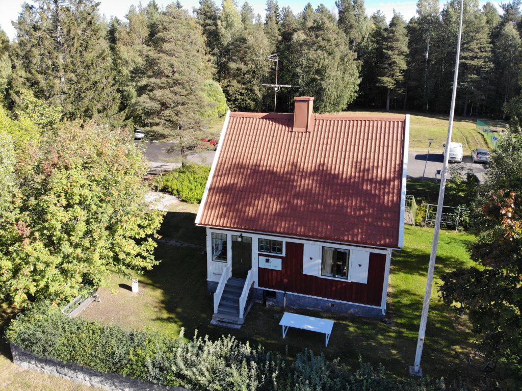 Ett litet rött trähus på en backe. Huset har fina snickardetaljer. Utanför huset syns en flaggstång. Huset är inramat av en häckplantering och höga träd.