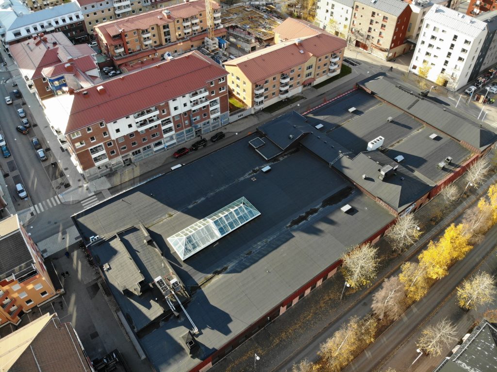 Översiktsbild fotograferad med hjälp av drönare. Bilden visar en småstad, byggnader och transportvägar. I fokus är en stor fastighet vars tak har ett glasparti inbyggt. Det är soligt och klart väder. Träden är färgade i höstgula toner.