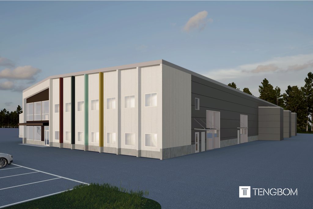 En grå industrihall står omgiven av tät barrskog. Byggnaden är enkel med plåtfasad och stora portar. Runtom syns asfalt och höga träd.