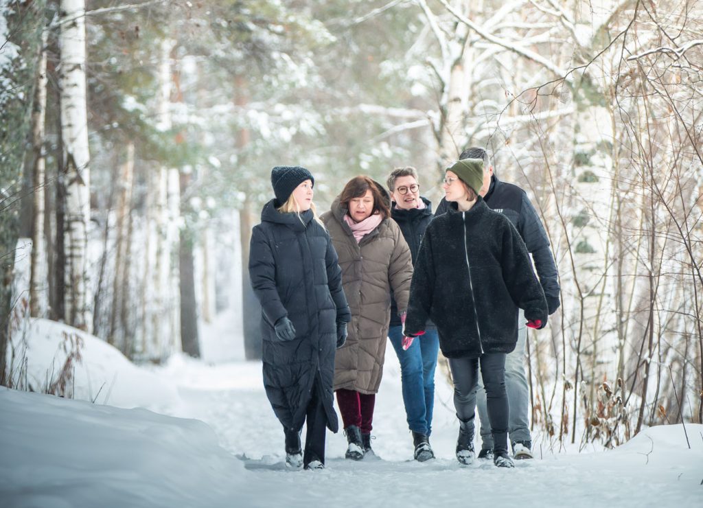 Fem personer går långsamt längs en smal stig i en snötäckt skog. Träden är täckta med ett tjockt lager vit snö som hänger från grenarna. Personerna är klädda i tjocka vinterkläder – jackor, mössor och vantar.