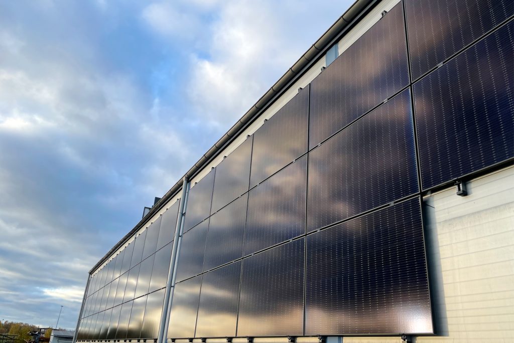 Bild på en byggnad med fasadhängda solcellspaneler. Panelerna är svarta och täcker en del av byggnadens fasad. De är monterade vertikalt längs väggen, vilket ger byggnaden ett modernt och hållbart utseende. Solcellspanelerna är jämnt placerade och reflekterar lätt ljuset från solen.