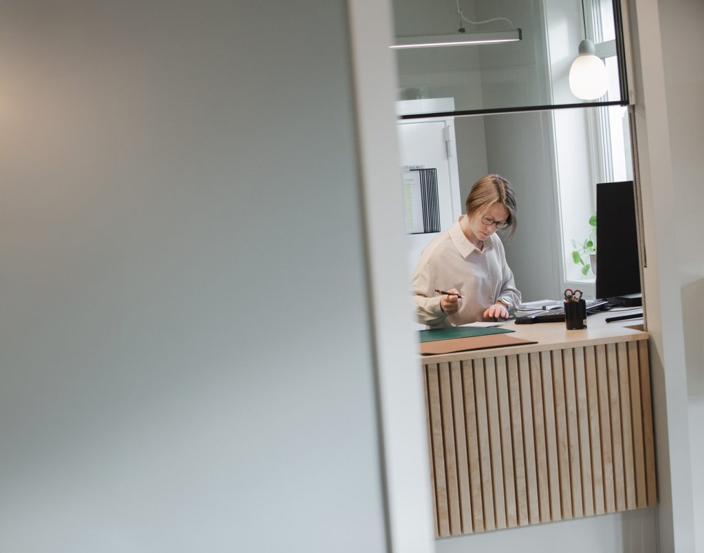 En kvinna står vid en receptionsdisk och antecknar. Hon ser koncentrerad ut. Framför henne syns en datorskärm och ovanför henne hänger en lampa.