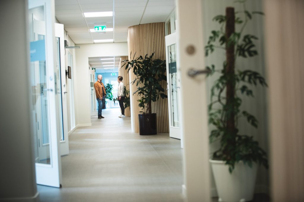 En man och en kvinna står i en korridor och samtalar. Mannen har en orangefärgad skjorta. Några dörrar i korridoren står på glänt. I förgrunden, på höger sida, syns en växt.