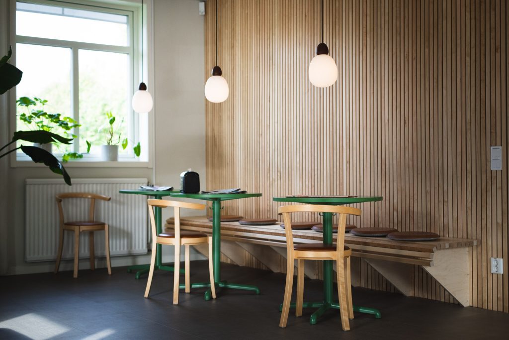 Interiörbild av ett fikarum. Väggen består av stående träpanel. Tre grönmålade bord står uppradade. Närmast borden finns en sittbänk av trä och ovanför borden hänger tre lampor.