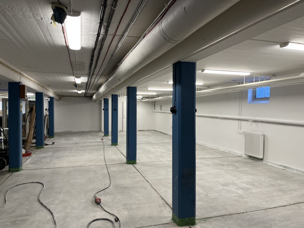 Interiörbild av en källare med vitt tak och betonggolv. I lokalen syns pelare som är målade i blått.