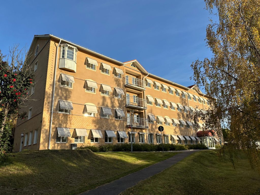 En gul tegelbyggnad med delvis putsad fasad står på en kulle. Solen lyser på byggnaden i ett varmt sken. På båda sidor syns björkar med höstfärgat lövverk.
