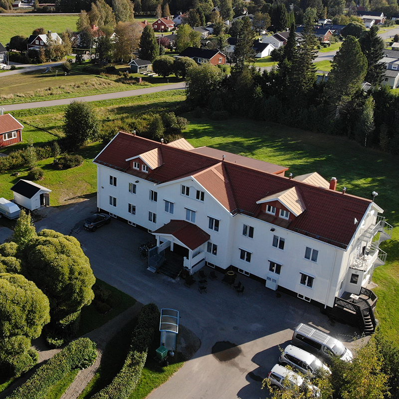 Översiktsbild av ett stort vitt hus, omgivet av parkeringsytor och transportvägar. Omgivningen är lummig och grön.