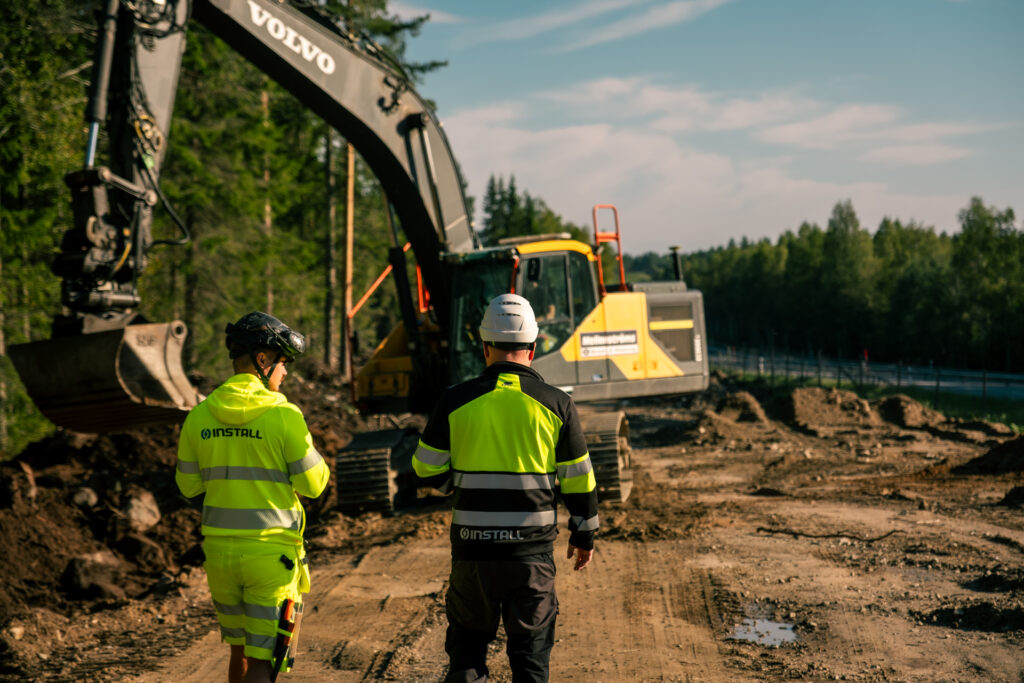 Två män går längs en lerig väg, klädda i varselkläder för arbete utomhus. I bakgrunden syns en grävmaskin och en klarblå himmel breder ut sig ovanför dem.
