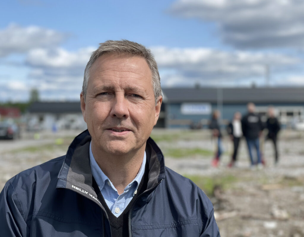 En man står på en industritomt. Han tittar in i kameran och ser nöjd ut. Ovanför är molnen både vit och mörkgrå. En grupp med människor står i bakgrunden och pratar.