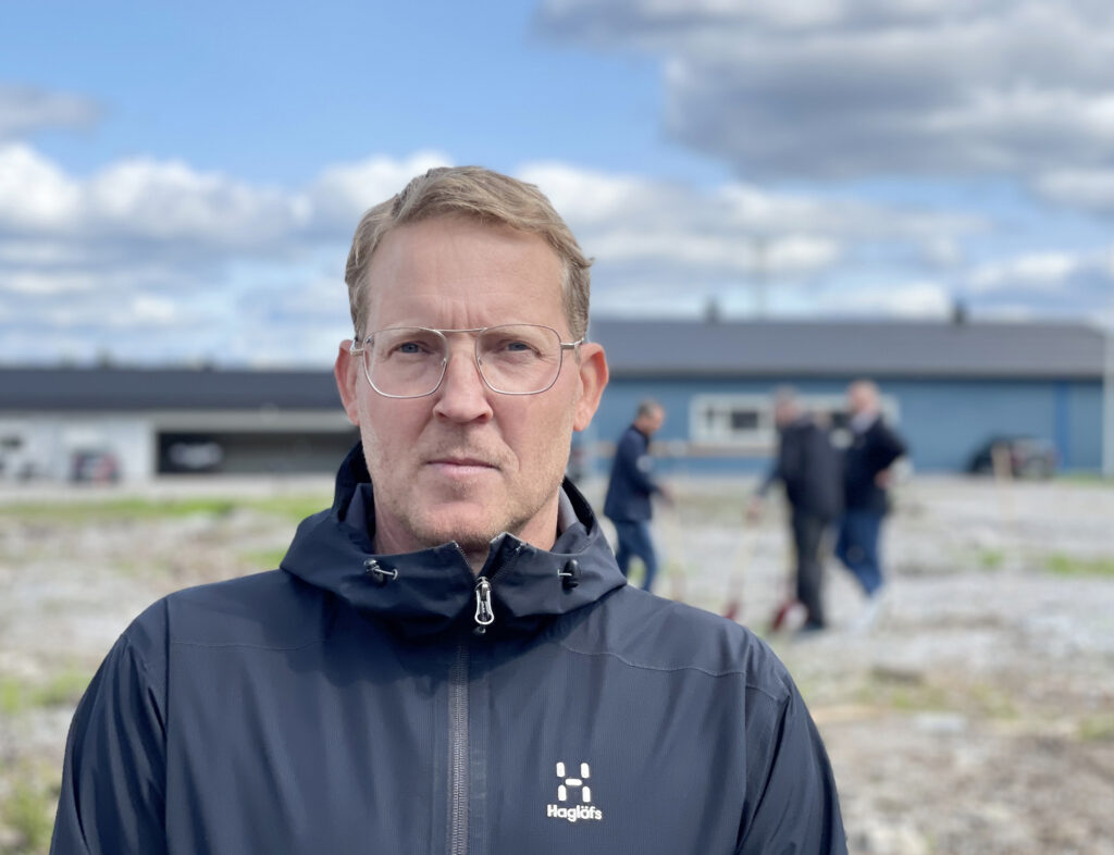 En man med mörkblont hår står på en industritomt. Han har glasögon med tunna bågar. Han tittar in i kameran och ser lugn ut.