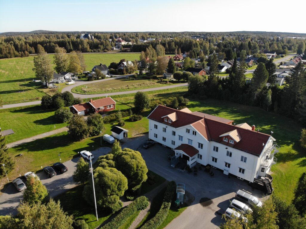 Bilden visar ett stort vitt hus i en lantlig och charmig miljö. Huset är omgivet av åkermark och transportvägar som leder fram till och runt byggnaden. Ett par fordon, bland annat en mindre lastbil och en personbil, står parkerade i närheten. I bakgrunden syns skog och berg som ramar in landskapet. Ljuset är mjukt och naturligt, och bilden förmedlar en stillsam och lantlig stämning.