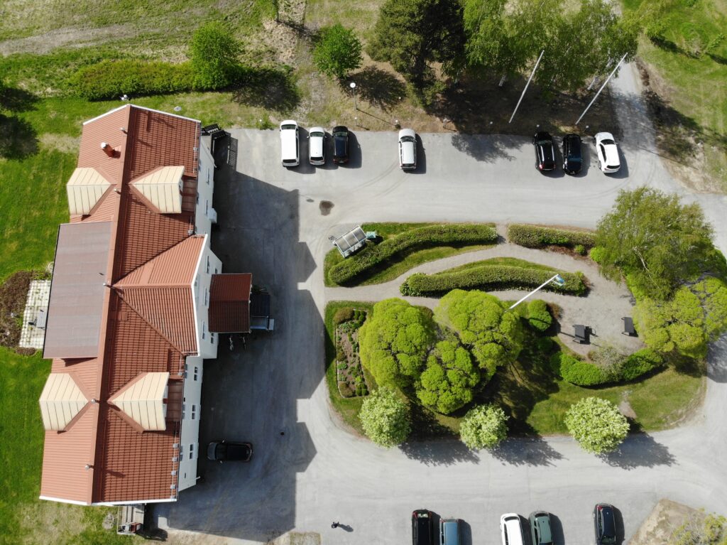 Drönarfoto taget rakt ovanifrån. Bilden visar det röda tegeltaket på en stor, vit byggnad samt en stor innergård med buskage och gångvägar i formationer. På parkeringen står flera bilar parkerade.