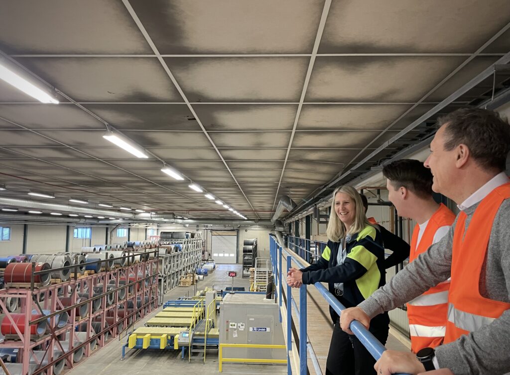 Två män och en kvinna står på en upphöjd sektion och samtalar. De befinner sig i en industriell miljö med högt i tak. I bakgrunden syns den rymliga industrihallen med synliga stålkonstruktioner och teknisk utrustning. Stämningen är glad, professionell och fokuserad.