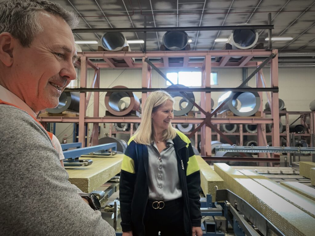 En man och en kvinna befinner sig i en industriell miljö med högt i tak. I bakgrunden syns den rymliga industrihallen med synliga stålkonstruktioner och teknisk utrustning. Stämningen är professionell och fokuserad.