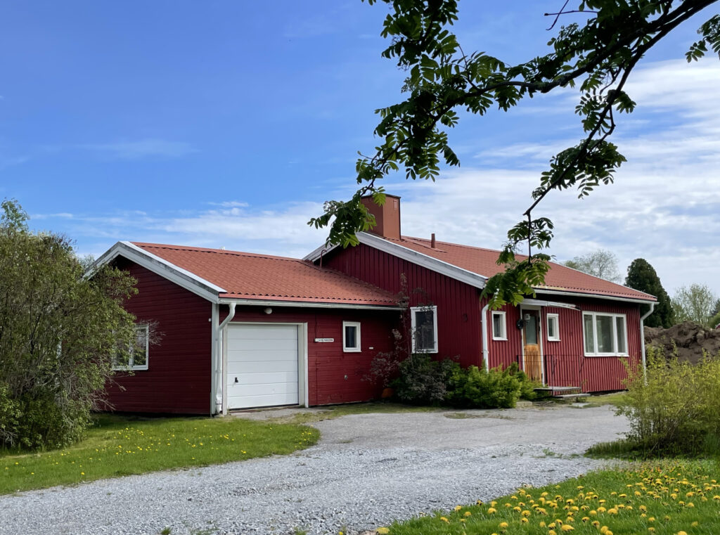 Ett litet rött hus med rött tegeltak står i en grön och somrig miljö. Taket är lätt sluttande, och huset omges av gräs och några utspridda träd. På ena gaveln finns en tillbyggnad i form av ett garage, målat i samma röda färg och med matchande tegeltak. Garaget smälter in i husets uttryck och förstärker det sammanhängande, lantliga intrycket. Ljuset är varmt och mjukt, och hela scenen utstrålar rofylld sommaridyll. Inga människor eller fordon syns i bilden.