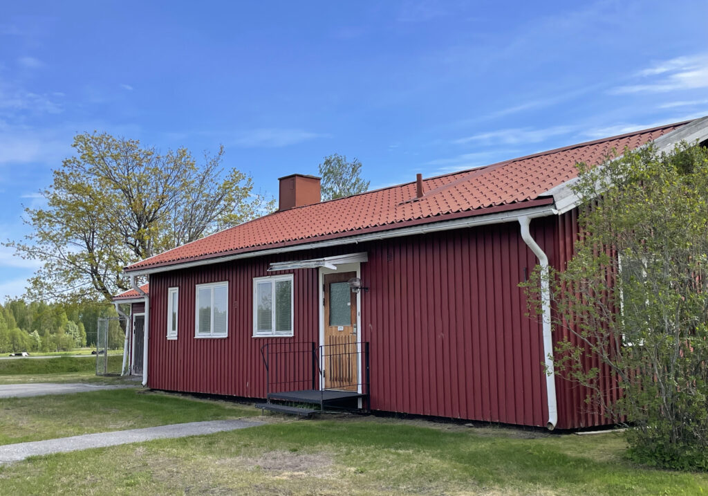 Ett litet rött hus med rött tegeltak står i en grön och somrig miljö. Taket är lätt sluttande, och huset omges av gräs och några utspridda träd.