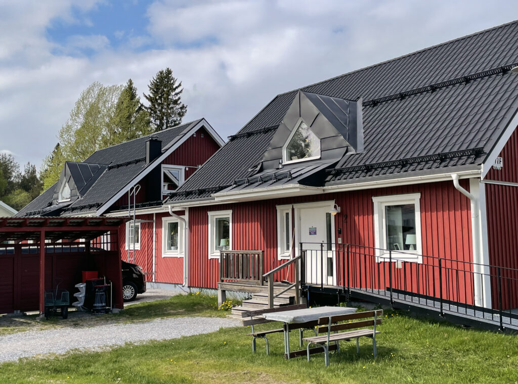 Ett rött parhus omgivet med en parkering i förgrunden. Himlen är lätt molnig. Träden i bakgrunden är lummigt gröna. Ett av husen har en ramp för rullstolsburna.