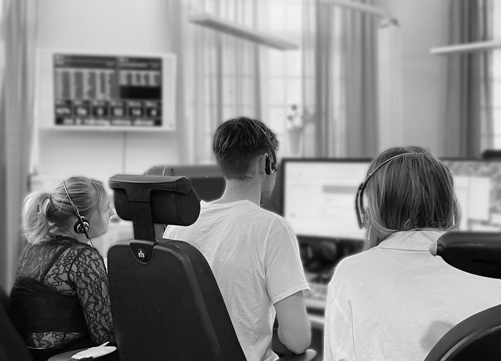 En svartvit bild som visar hur tre personer, en man och två kvinnor, sitter framför en datorskärm. Bilden är tagen bakifrån och visar ryggarna på samtliga personer. Varje person på bilden har på sig headset. Bilden ger en dokumentär känsla.