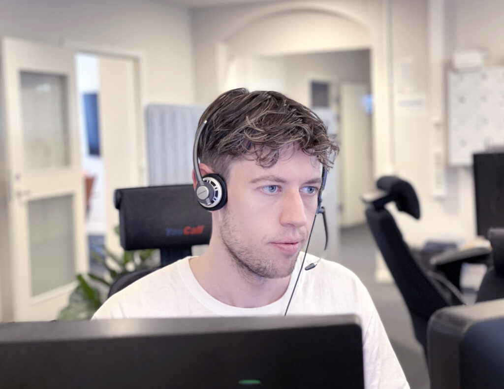 Bilden är en porträttbild på en operatör. På huvudet har han ett headset med hörlurar och liten mikrofon närmast munnen. Han sitter framför en datorskärm. Hans blick är fokuserad på skärmen. Han ser koncentrerad ut.