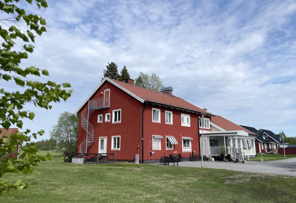 Ett rött tvåvåningshus med vita knutar står bredvid en asfalterad infart. Huset har ett traditionellt sadeltak och är förlängt med en tillbyggnad i samma stil men i en ljus fasadfärg, vilket ger ett harmoniskt och sammanhållet intryck. Bilden förmedlar en stillsam och naturnära känsla, med mjukt dagsljus över landskapet.