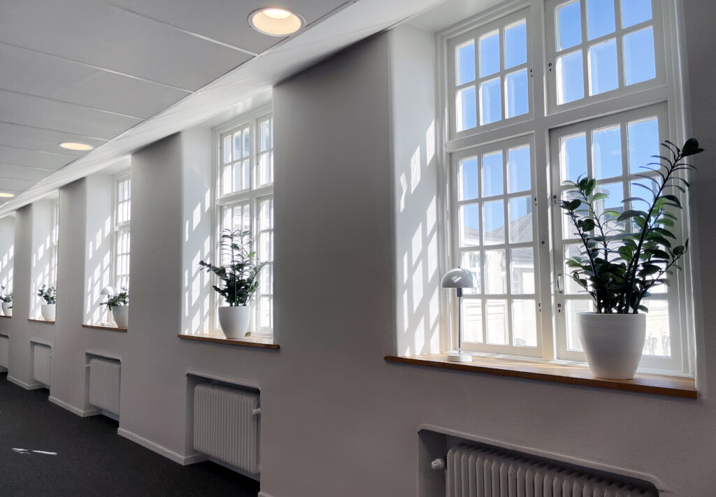 En ljus hall med stora, eleganta fönster som släpper in rikligt med dagsljus. Ljuset spelar mot ljusa väggar och golv i en stil som kombinerar den äldre byggnadens karaktär med moderna detaljer. Utrymmet är luftigt och tillgängligt, med god framkomlighet och stilren inredning som speglar en nutida arbetsmiljö i historisk miljö.