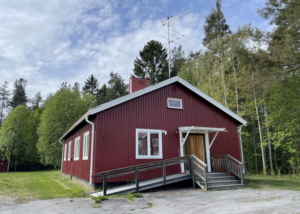 Ett litet rött hus står omgivet av gräs och lummiga träd. Huset har vita knutar och ett sadeltak, vilket ger ett traditionellt och hemtrevligt intryck. Grönskan runt huset är tät, och inga andra byggnader eller fordon syns i bilden. Ljuset är mjukt och förstärker den stilla, naturnära känslan.