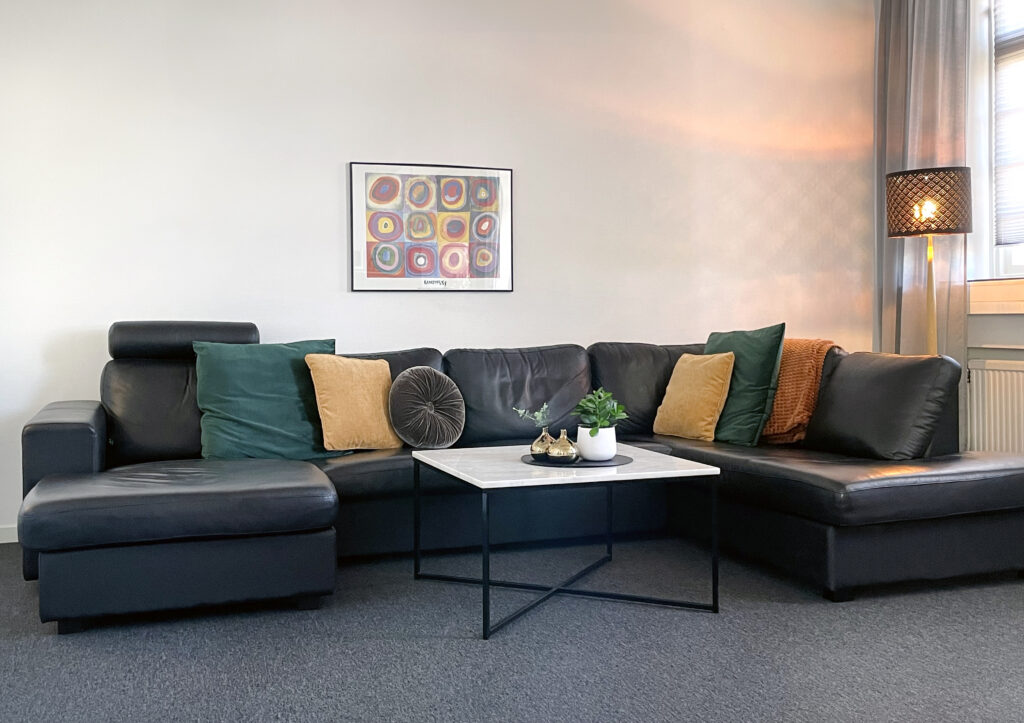 Bilden visar interiören från ett socialt rum där man kan slappna av men samtidigt vara social. I mitten av rummet står en mörkgrå soffa. I soffan finns kuddar med olika kulörer på kuddfodralen. Till höger om soffan står en lampa som lyser i mjukt sken.