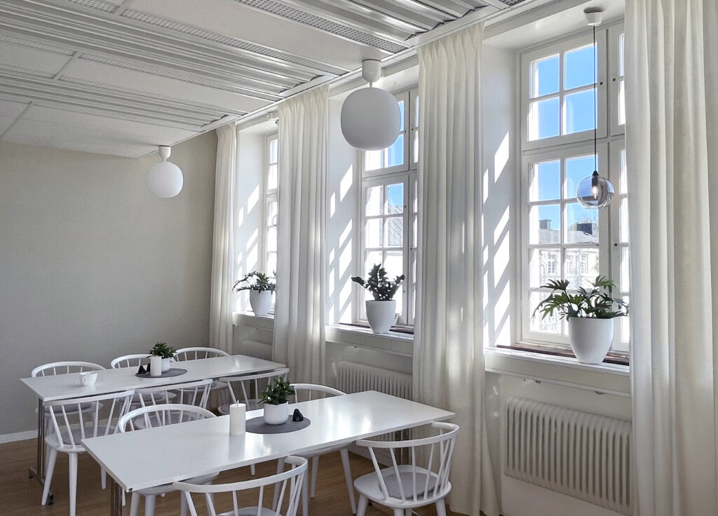 Bilden visar en lunchrum i framförallt vit kulör. Två bord står närmast de höga fönstren till höger. Solen lyser in, varmt och mycket. I varje fönster står en grön krukväxt. Bilden är harmonisk och lugn.