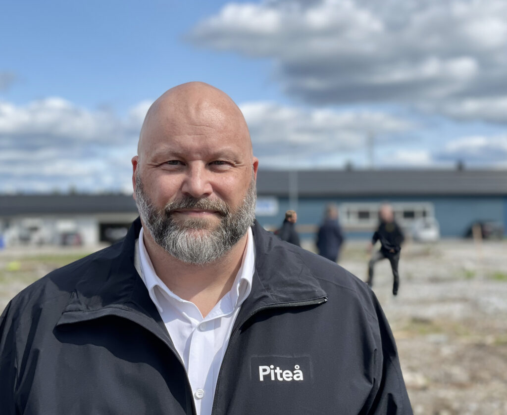 En man med skägg står framför kameran. Han ser lugn och glad ut. Hans blick är klar. Han har på sig en mörkblå skaljacka med logotypen Piteå på vänster sida av jackan. I bakgrunden står en grupp människor och pratar.