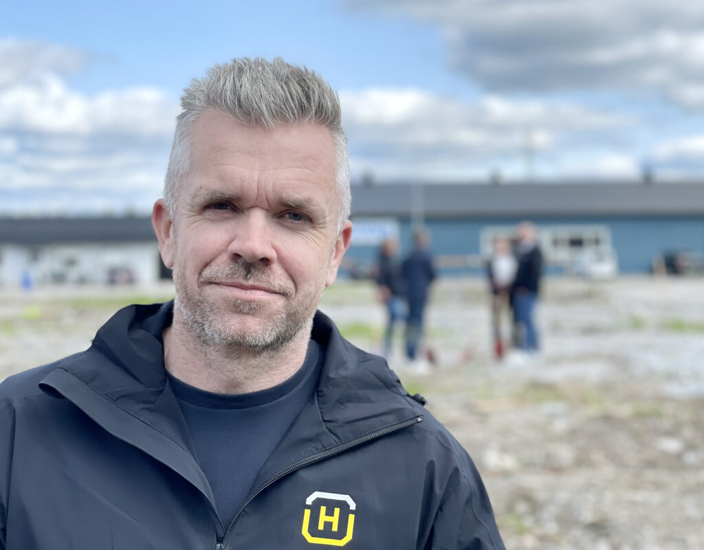 En man står framför kameran. Han tittar rakt in i kameran och ser nöjd och lugn ut. Han har på sig en t-shirt och ovanpå en mörkblå skaljacka. På vänster sida av jackan syns en logotyp i form av ett H, som står för Hellströms bygg. Himlen består av både vita och mörkgrå moln.