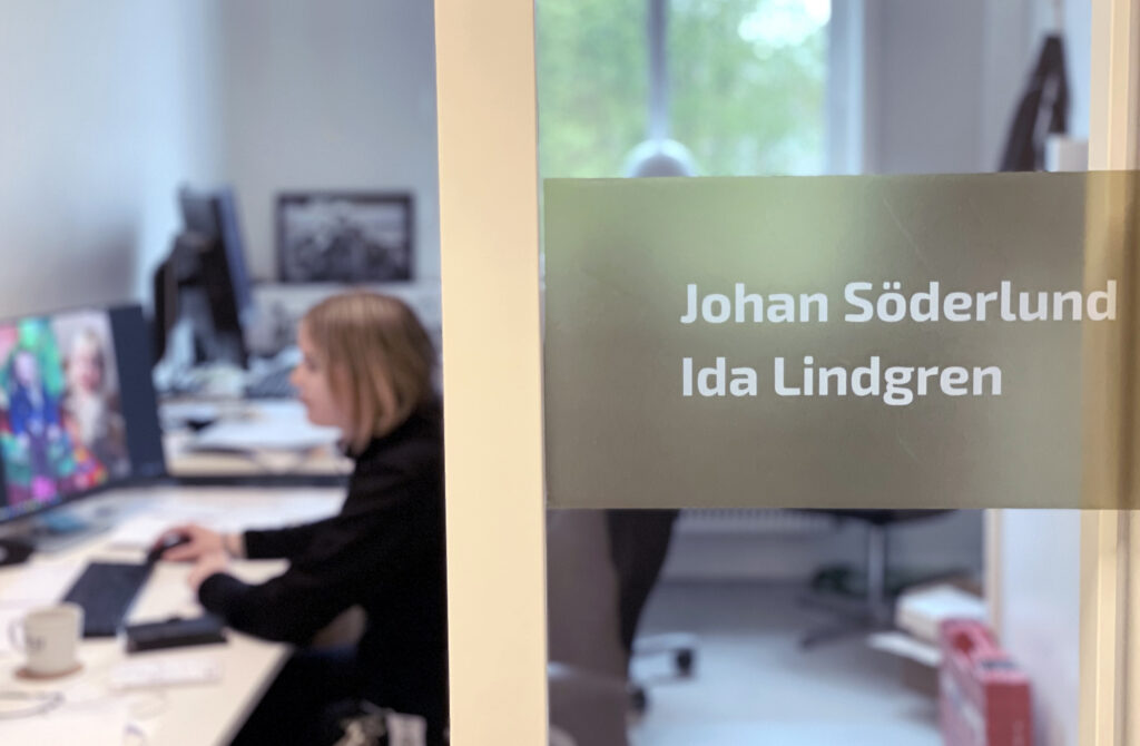 Bilden visar ett kontor. I bakgrunden, som är något oskarp, sitter en kvinna och arbetar vid datorn. Bildens fokus är två namnskyltar på glasdörren som informerar om vilka som sitter i kontoret, nämligen Johan Söderlund och Ida Lindgren.