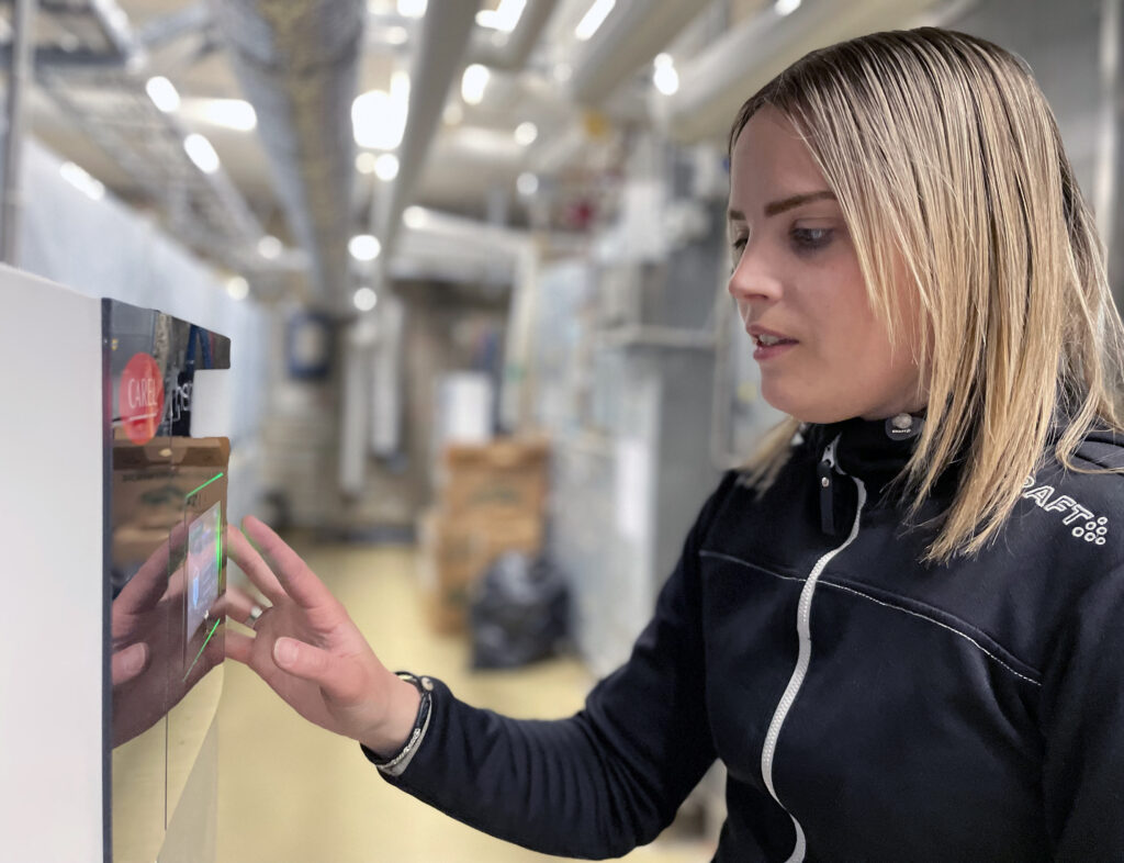 Bilden visar en närbild på en kvinna som står i ett fläktrum. Hon gör inställningar på en luftfuktare som förser ventilationen med luft. Kvinnan ser koncentrerad och fokuserad ut. Hon har sin ena hand på luftfuktarens display.
