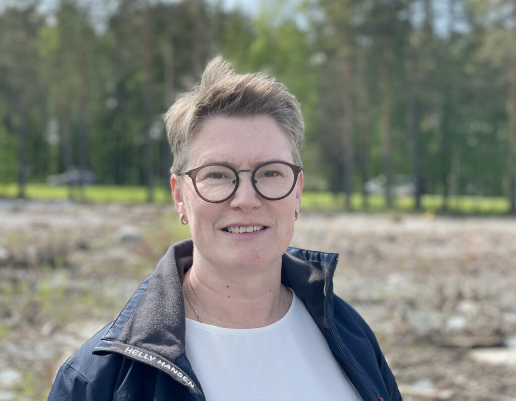 En kvinna med kortklipp frisyr tittar rakt in i kameran. Hon ler lite lätt och ser nöjd och säker ut. Hon har på sig glasögon med mörkbruna bågar. I bakgrunden syns skog.