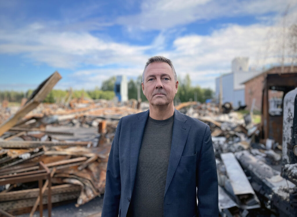 En man står framför en nedbrunnen fastighet. Han är ledigt men propert klädd och har ett allvarligt, men inte argt, uttryck i ansiktet. Ovanför syns en blå himmel med vita moln.