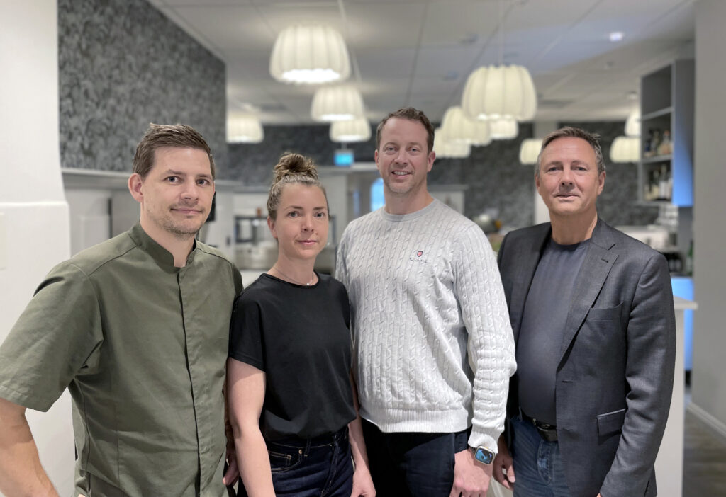 Ett gruppfoto med fem personer som står tillsammans i en restaurangmiljö. Alla tittar in i kameran, leende och nöjda. De är lätt uppklädda och bilden har en varm känsla.