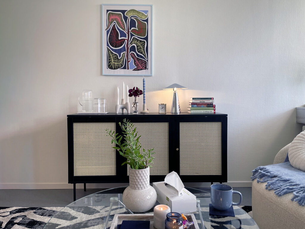 Interiörbild från mottagningsrummet. I fokus står en byrå med böcker, ljus och en lampa ovanpå. I förgrunden syns ett glasbord. På bordet står en vas med gröna kvistar i. Bilden är varm och behaglig.