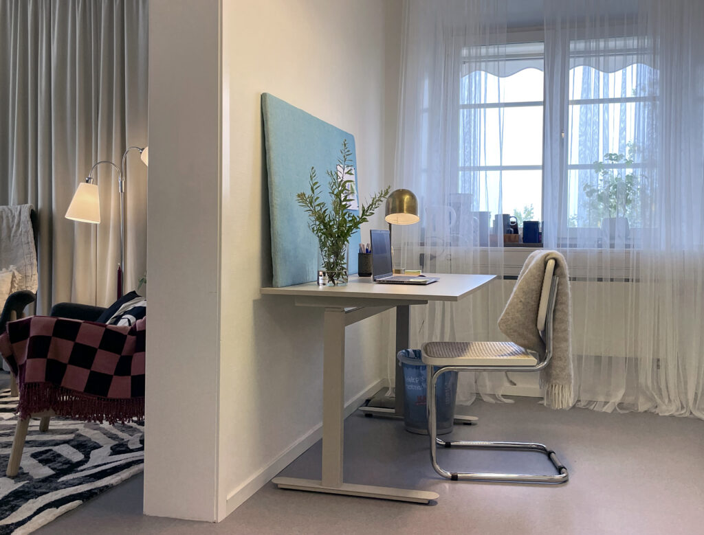 Bild som visar indelningen i mottagningen. Till höger syns en arbetsstation med skrivbord, dator, laptop och en vas med gröna kvistar. Till vänster i bilden skymtar mottagningsrummet med fåtöljer.