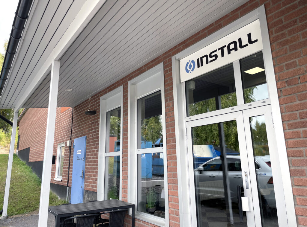 Byggnad med tegelfasad. Stora fönster och en glasdörr pryder entrén, ovanför sitter en vit skylt med svarta bokstäver som bildar ordet Install.