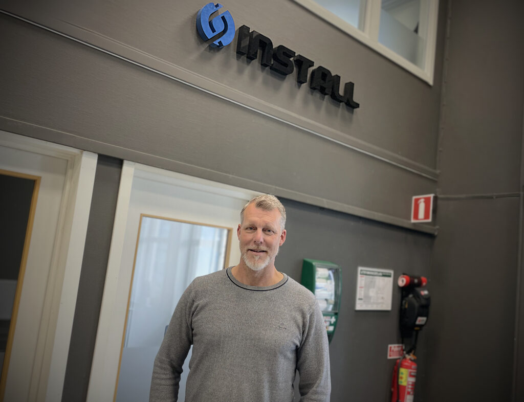 Porträtt av en man med lätt skäggväxt, fotad snett underifrån. Han ler och ser nöjd ut. Ovanför honom syns en skylt med svarta bokstäver som bildar ordet Install. I bakgrunden, mot en grå vägg, sitter första hjälpen-utrustning.
