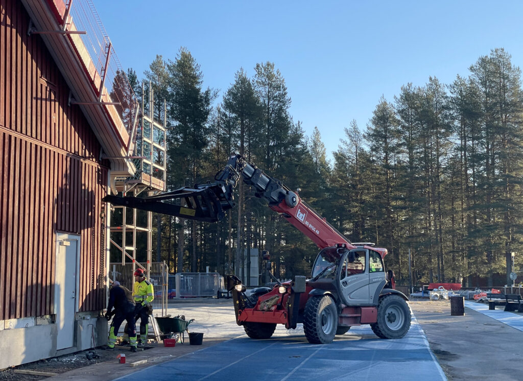 Två personer arbetar vid ställningarna längs fasaden under klar blå himmel. Ett arbetsfordon står intill byggnaden där renoveringen pågår.