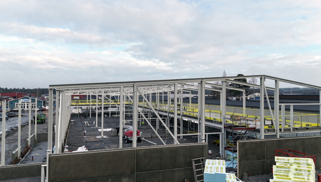 Drönarbild över byggnation av industrihall med synlig stomme och bjälklag. Grus i botten, mulen himmel och en tydlig, dokumentär känsla.