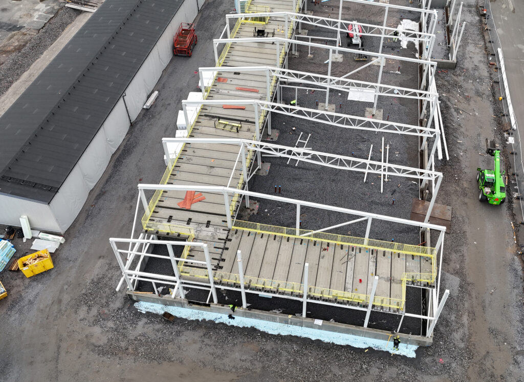 Drönarbild över byggnation av industrihall med synlig stomme och bjälklag. Grus i botten, mulen himmel och en tydlig, dokumentär känsla.