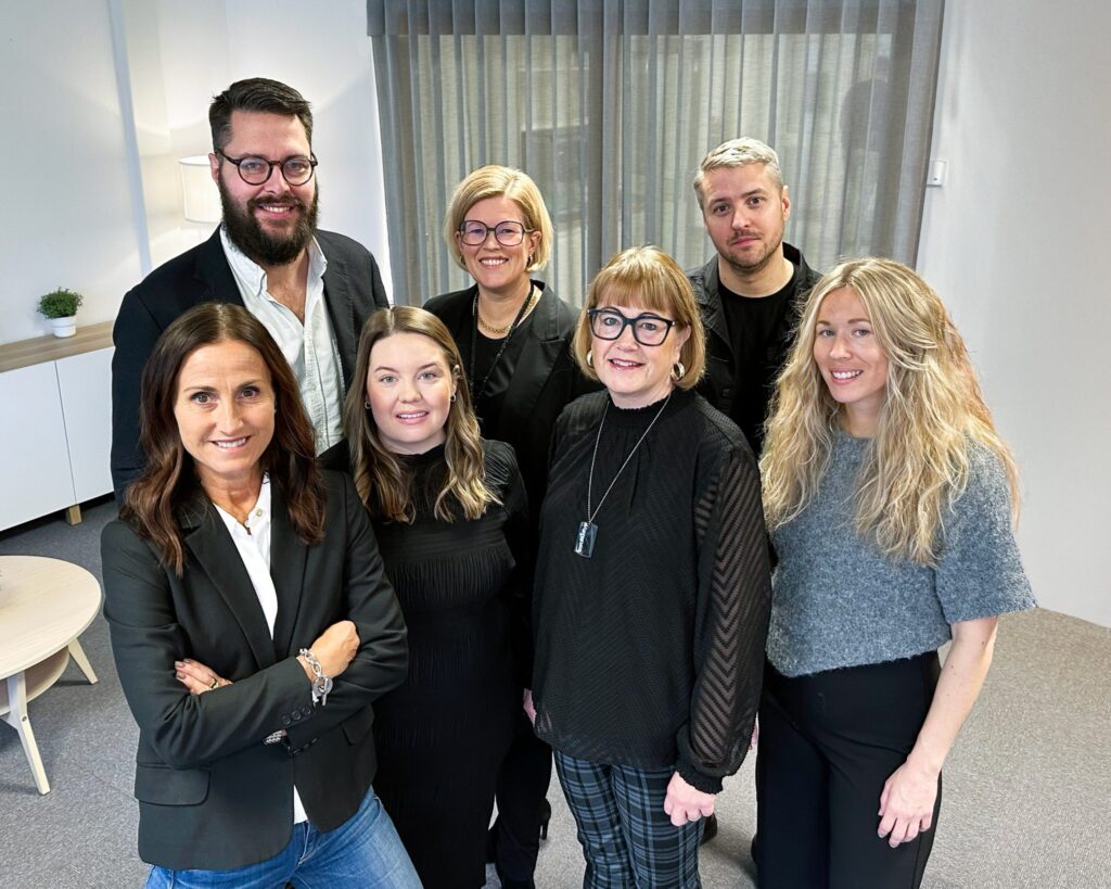 Gruppbild tagen lite ovanifrån med sju personer från inköp, två män och fem kvinnor i ledigt snygga kläder. De står samlat, ler mot kameran och ger ett lugnt och glatt intryck.