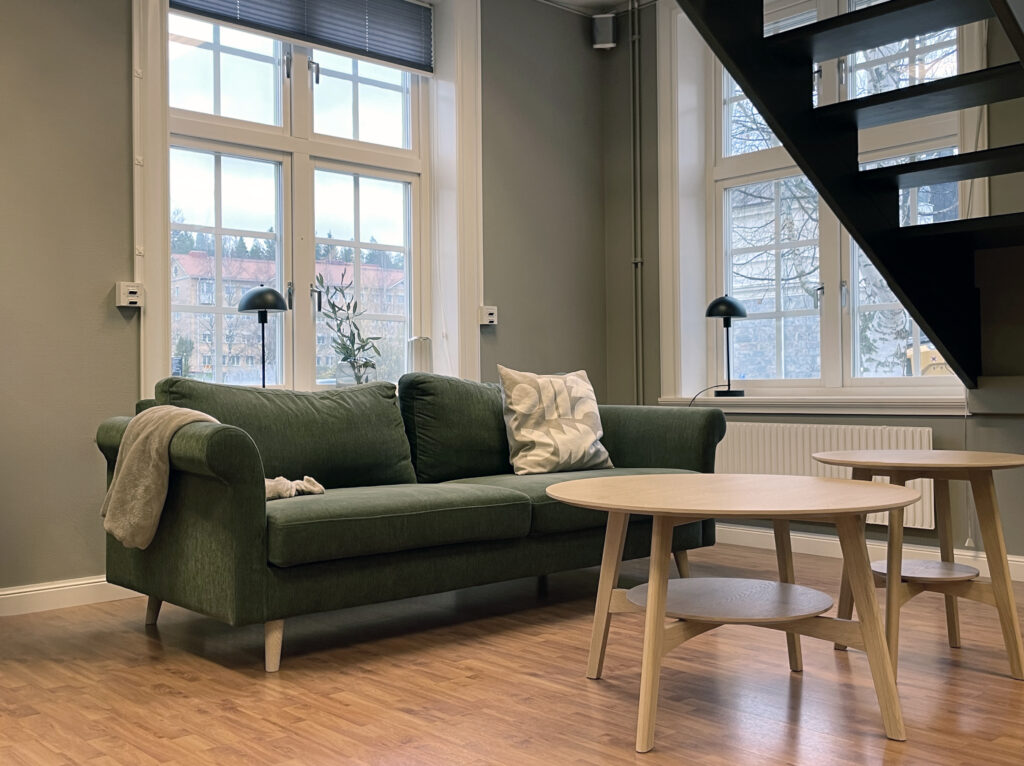 Ett kontor med lugna, sobra färger där mjuka detaljer skapar en harmonisk interiör. I hörnet står en soffa och i förgrunden syns en svart trappa.