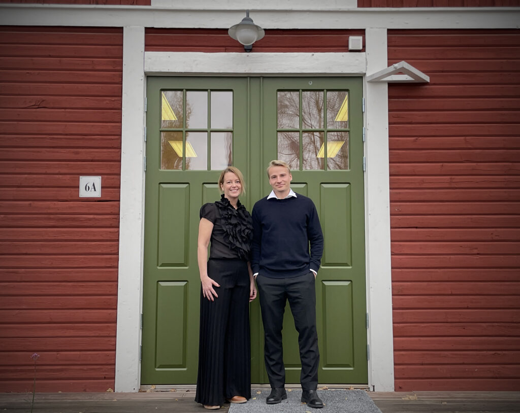 En kvinna och en man står framför en grön entrédörr mot röd träfasad och möter kameran med mjuka, nöjda leenden som ger bilden en varm och trygg känsla.