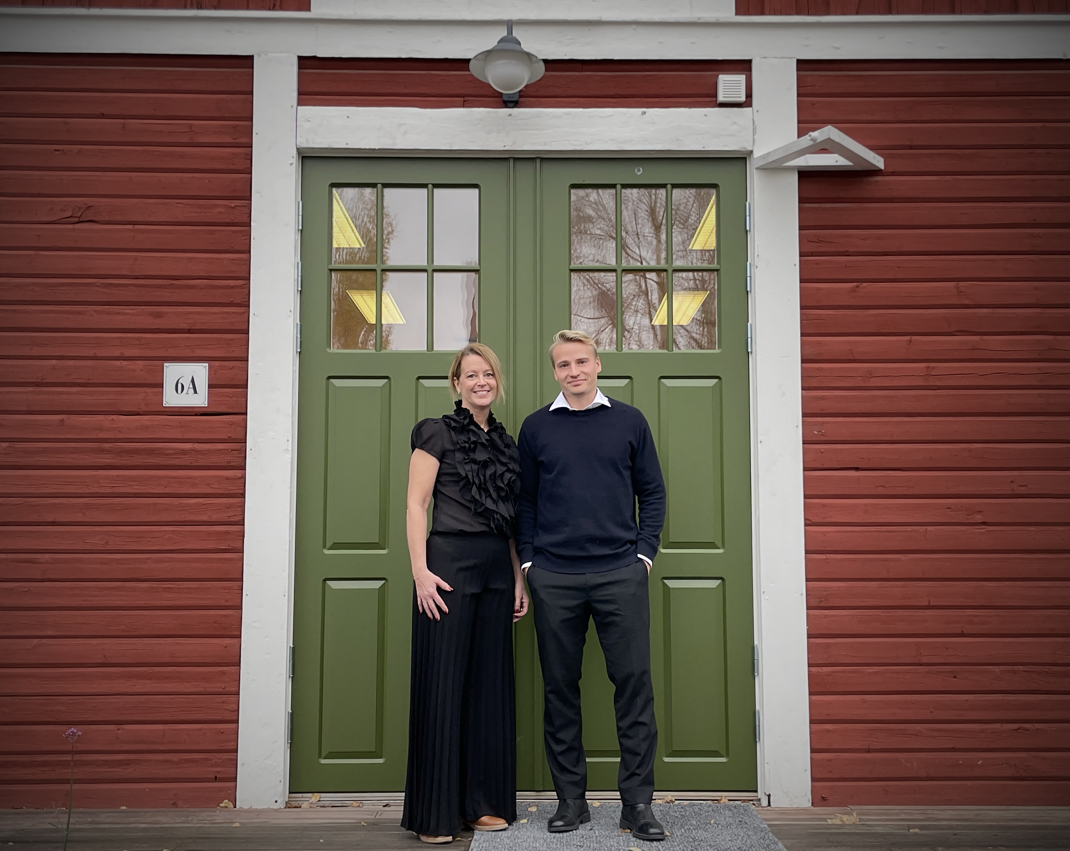 En kvinna och en man står framför en grön entrédörr mot röd träfasad och möter kameran med mjuka, nöjda leenden som ger bilden en varm och trygg känsla.