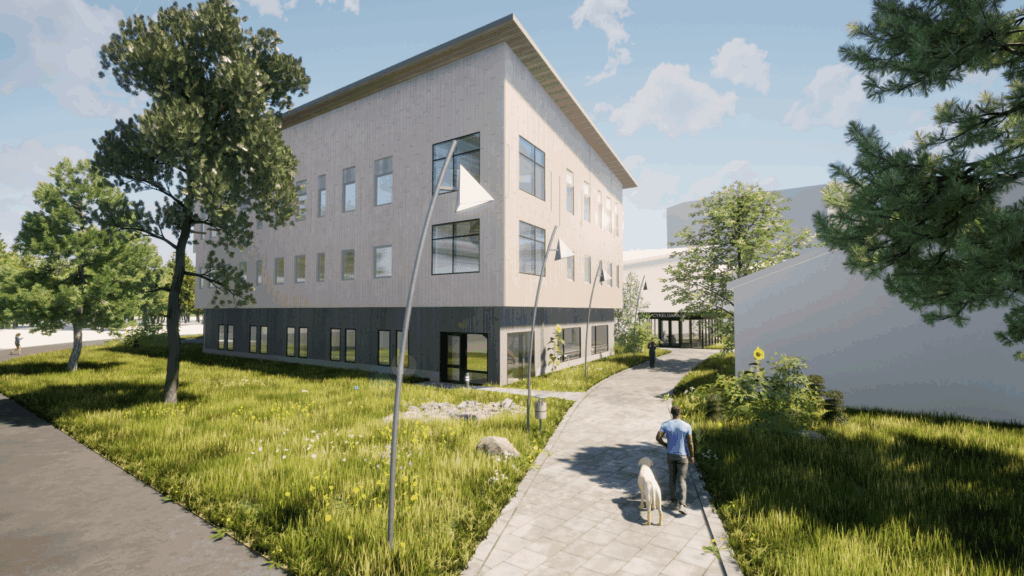 Ett stilrent kontorshus i tre våningar med varm träfasad omgivet av grönt och stående under en blå himmel med lätta moln som ger ett öppet och inbjudande intryck. En man med en hund går längs promenadstråket närmast i bild.